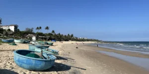 Blue fishing boat resting on golden sand at Mui Ne beach during low tide, with palm trees and the resort strip visible along the shoreline