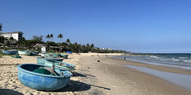 Blue fishing boat resting on golden sand at Mui Ne beach during low tide, with palm trees and the resort strip visible along the shoreline