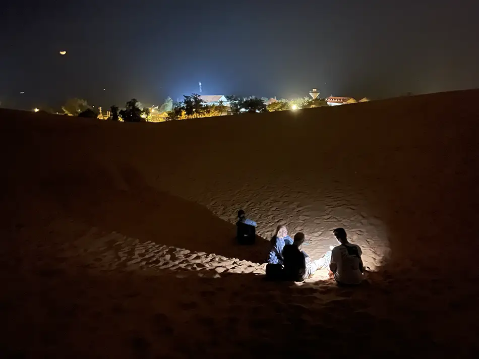 Travelers sitting on the red sand dunes at night with city lights and hotel glow visible along the coastline below