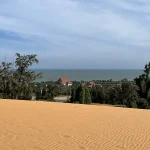 Wide view of the Mui Ne red sand dunes rising above the coastal road under a clear sky, with sparse trees along the base