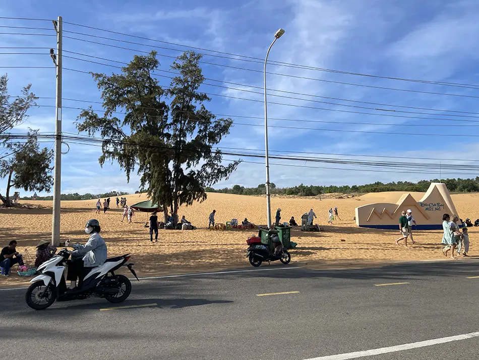 Visitors gathering on the sand near the road as late afternoon begins, with motorbikes parked along the entrance area