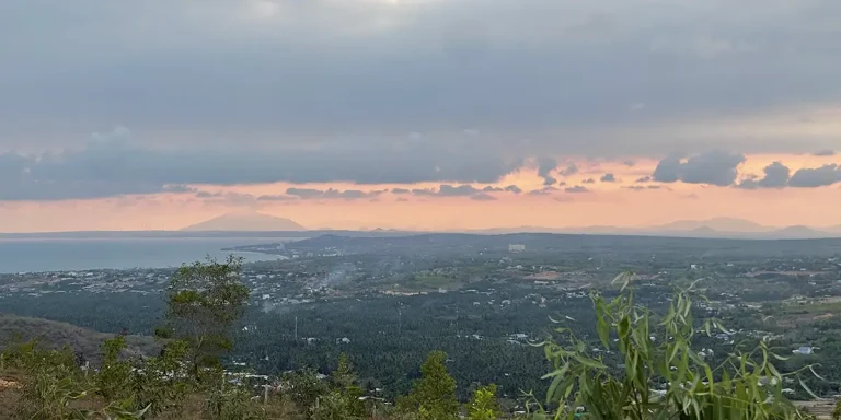 Sweeping panoramic view of Mui Ne's coastline and village rooftops seen from an elevated vantage point at dusk, with pink and purple clouds reflected on the calm sea — a Mui Ne hidden gem known to few visitors