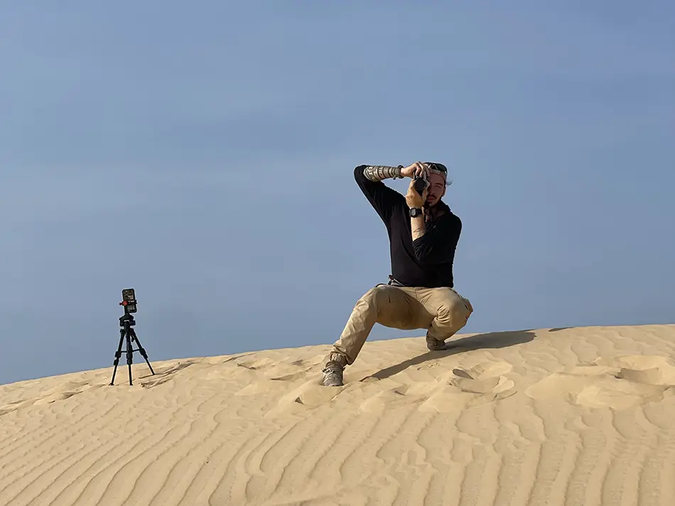 Traveler kneeling on a high sand dune to take a photo, with bright sky and flat terrain stretching behind