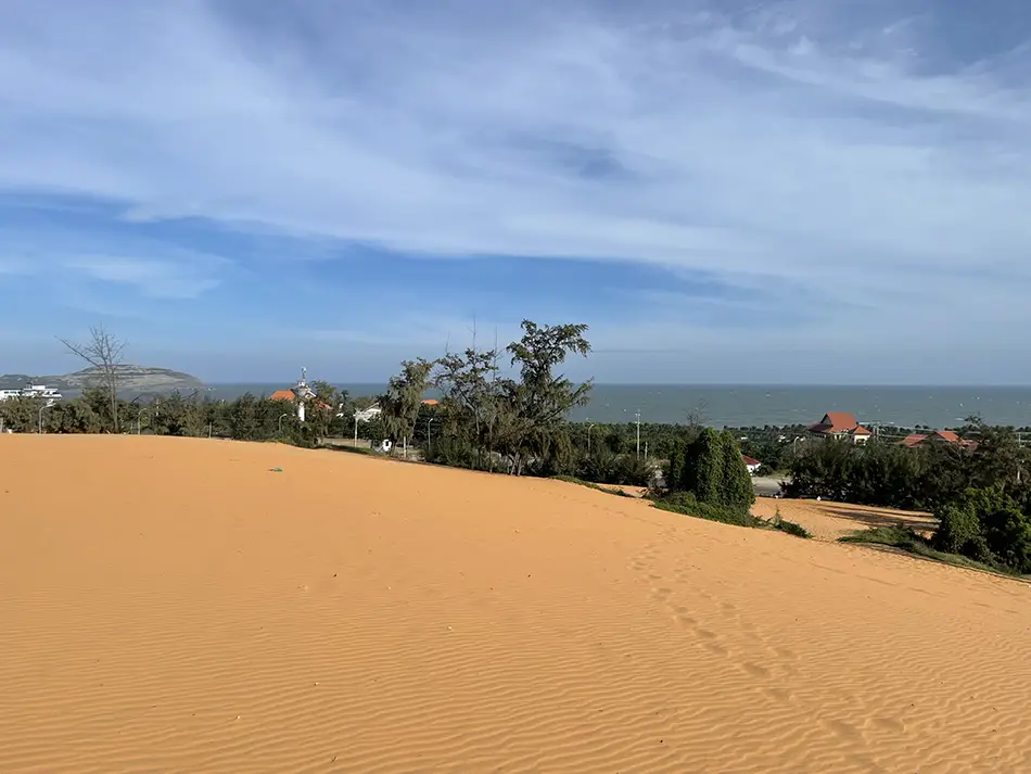Coastal panorama from the higher points of the red dunes showing vegetation, rooftops of the nearby village, and the sea beyond