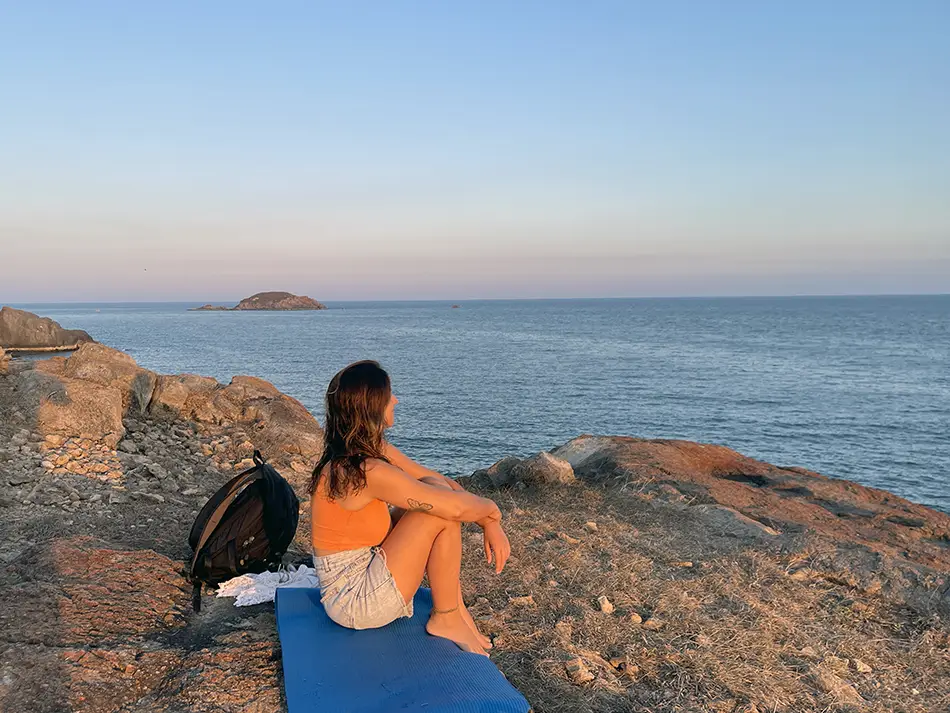 Young traveler in an orange top sitting on large granite boulders at a secluded rocky beach in Mui Ne, watching the sunset over the South China Sea