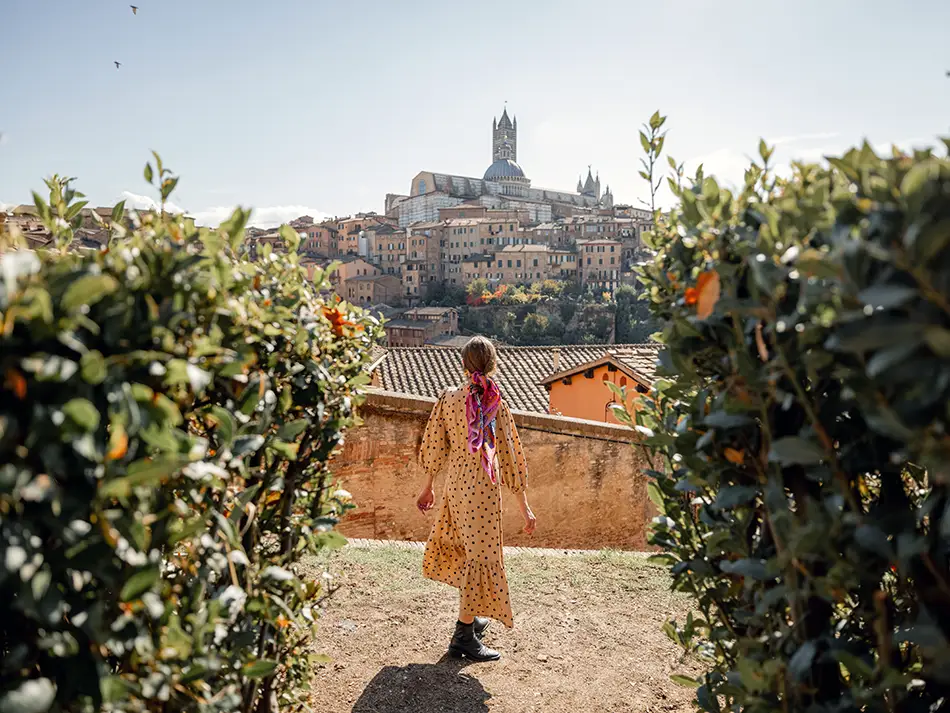 A woman in a red dress walks along a scenic hilltop path surrounded by foliage with the medieval towers and rooftops of Siena's old town visible in the Tuscan wine country