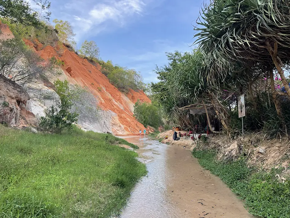 Wide view of the sculpted red and orange sand formations along Suoi Tien, with a shallow grey stream running through the base of the canyon