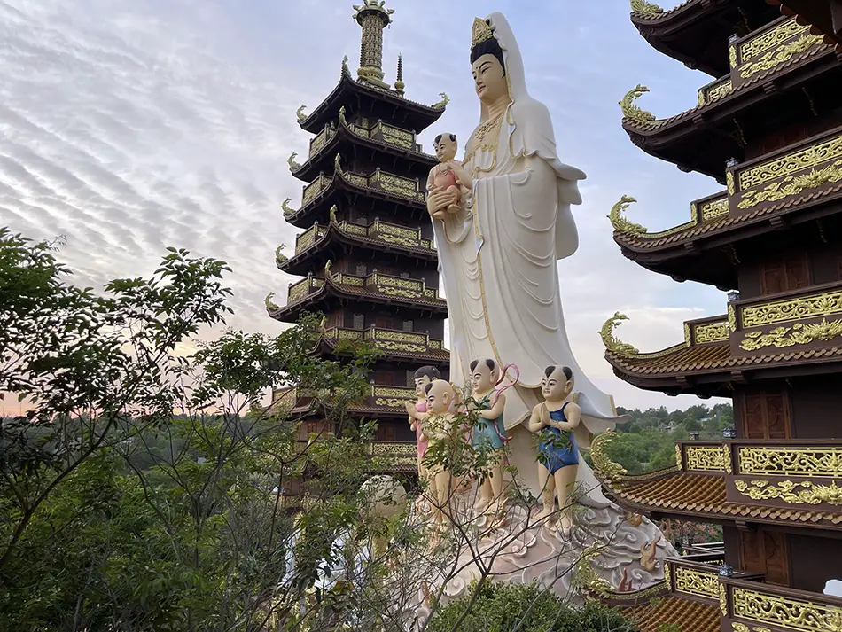 Multi-tiered Buddhist pagoda and large standing Guanyin statue at Thien Quang Temple in Mui Ne, set against a partly cloudy sky with green hillside in the background