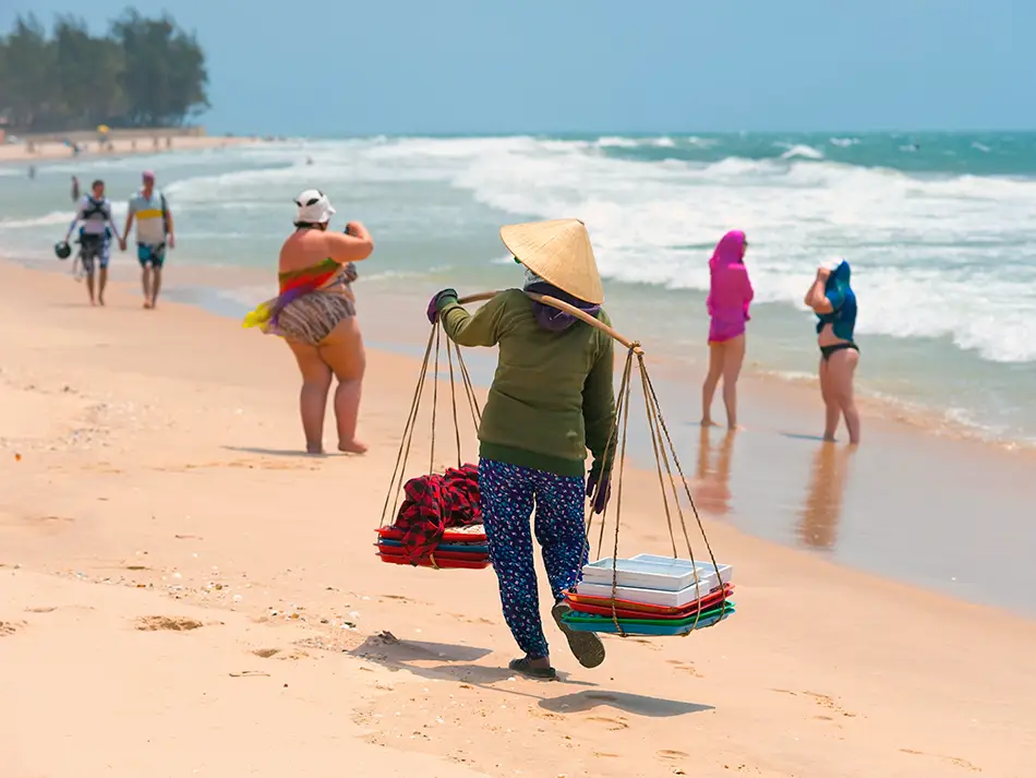 Elderly Vietnamese woman carrying goods in baskets on a shoulder pole, walking past tourists on the beach.