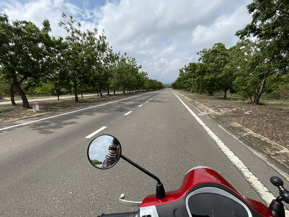 First-person view riding an electric scooter along a smooth paved road lined with trees in Vietnam.