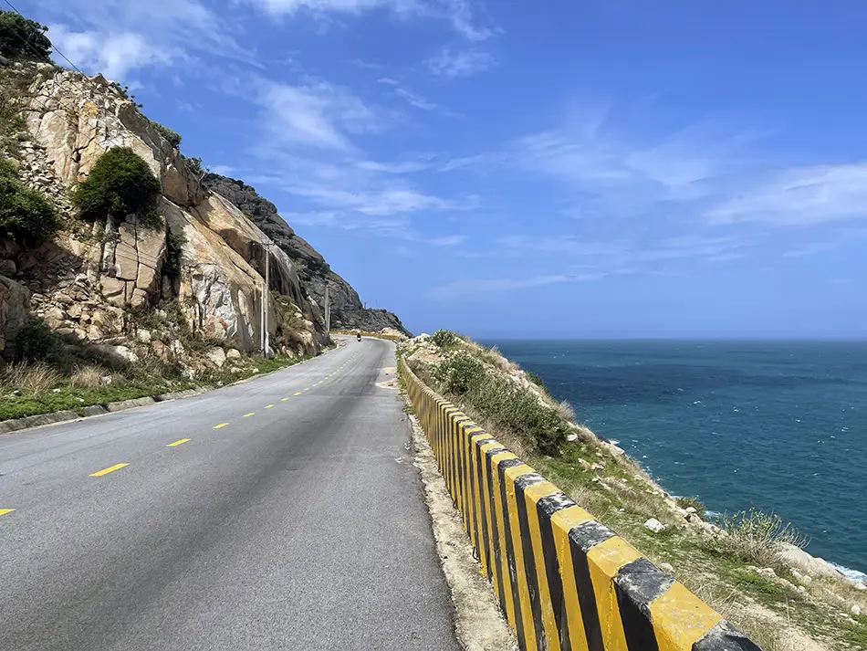 First-person driver view from a motorcycle on a coastal winding road overlooking the sea.