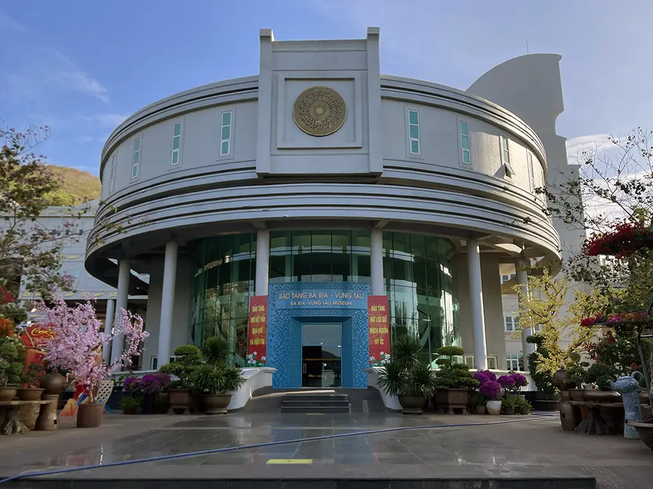 Modern teal-and-glass exterior of the Ba Ria–Vung Tau Provincial Museum, with trees and pink flowering plants in front of the curved facade.