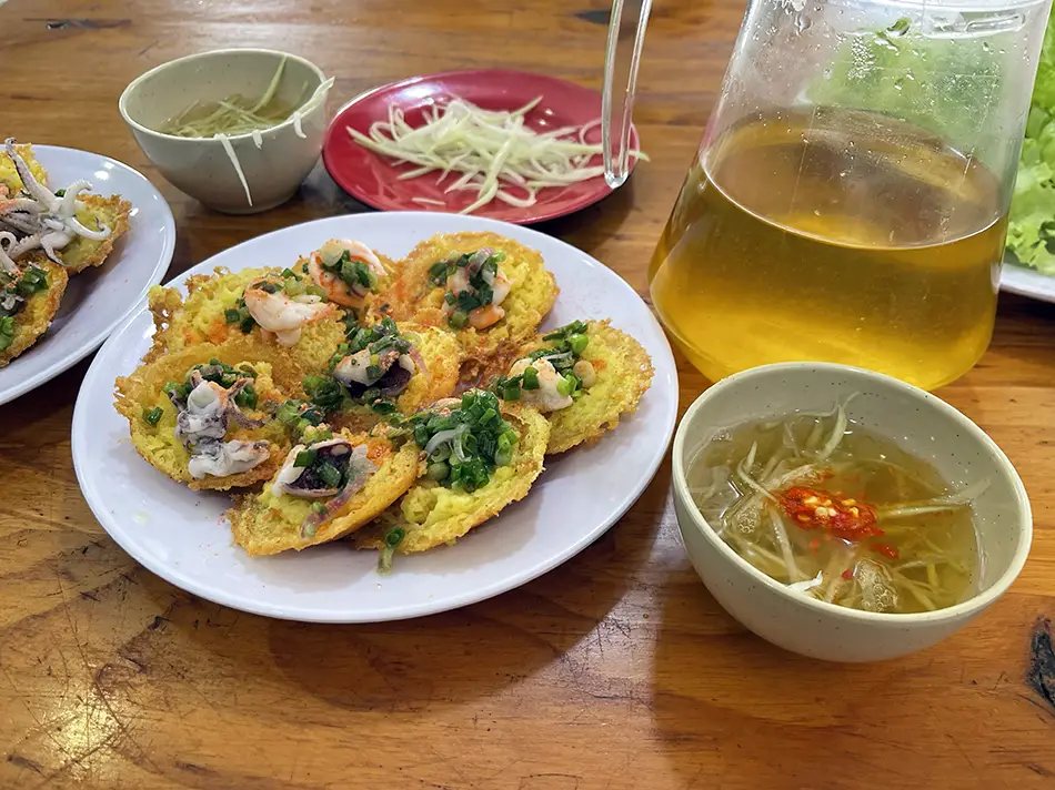 A plate of bánh khọt, small crispy rice pancakes topped with shrimp, served with fresh greens and a bowl of dipping sauce.