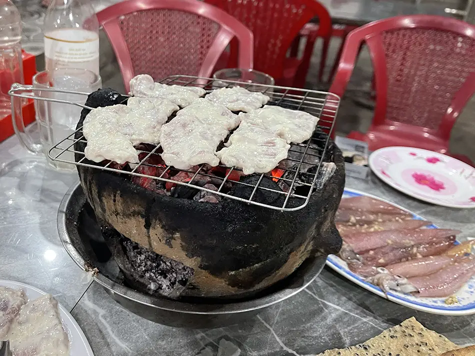Small charcoal grill on a Vietnamese bia hoi table with skewers cooking and raw meat plated alongside