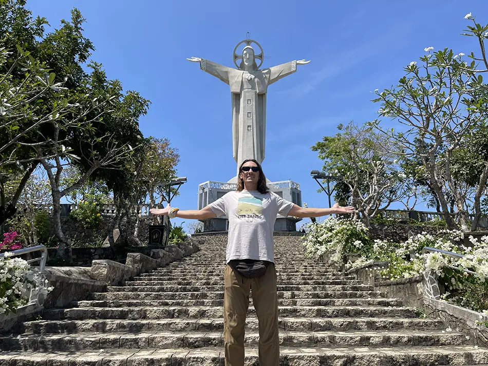 A visitor standing with arms outstretched at the base of the stone stairway leading up to Vung Tau's Christ the King Statue, flanked by tropical garden landscaping on a sunny day — one of the city's most iconic landmarks
