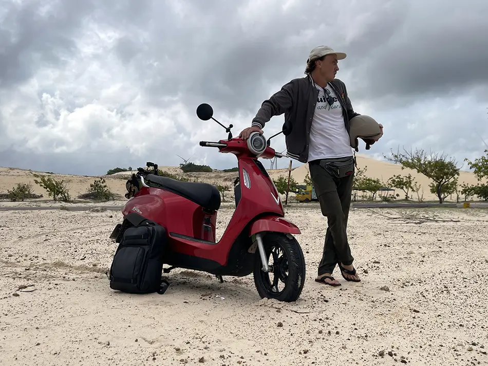 Yuri Malykh standing with his electric scooter against the background of white sand dunes.