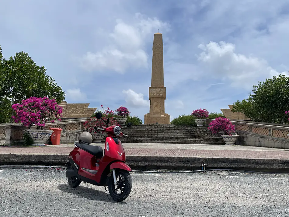 Red electric scooter parked beside a Ho Chi Minh Trail monument with flowers and a tall stone pillar