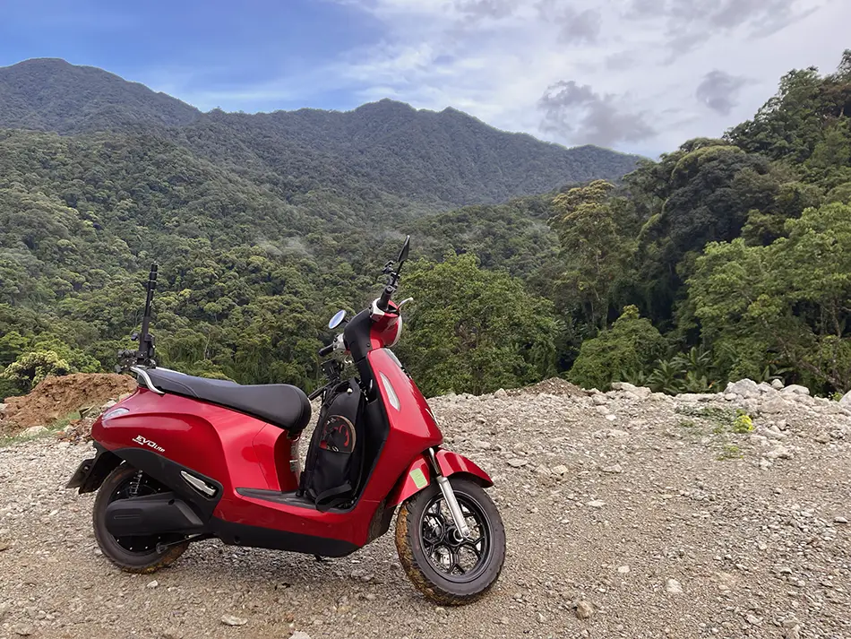 An electric scooter parked on the shoulder of a winding road overlooking wild dense vegetation.