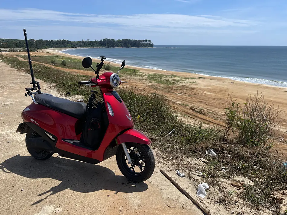 A red electric scooter requiring no driver's license parked on a beach in Vietnam surrounded by plastic litter.