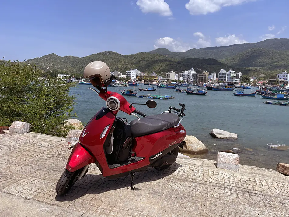 An electric scooter parked on a waterfront with boats in the harbor and a green mountain in the background.
