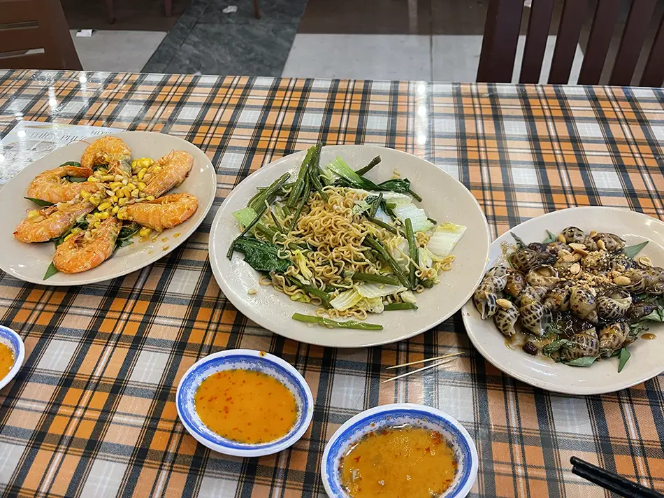 Multiple plates of Vietnamese seafood — including grilled whole fish, shellfish, and small bowls of dipping sauce — spread across a checkered tablecloth at a local restaurant; sampling fresh seafood is consistently highlighted among the best Vung Tau travel tips for first-time visitors