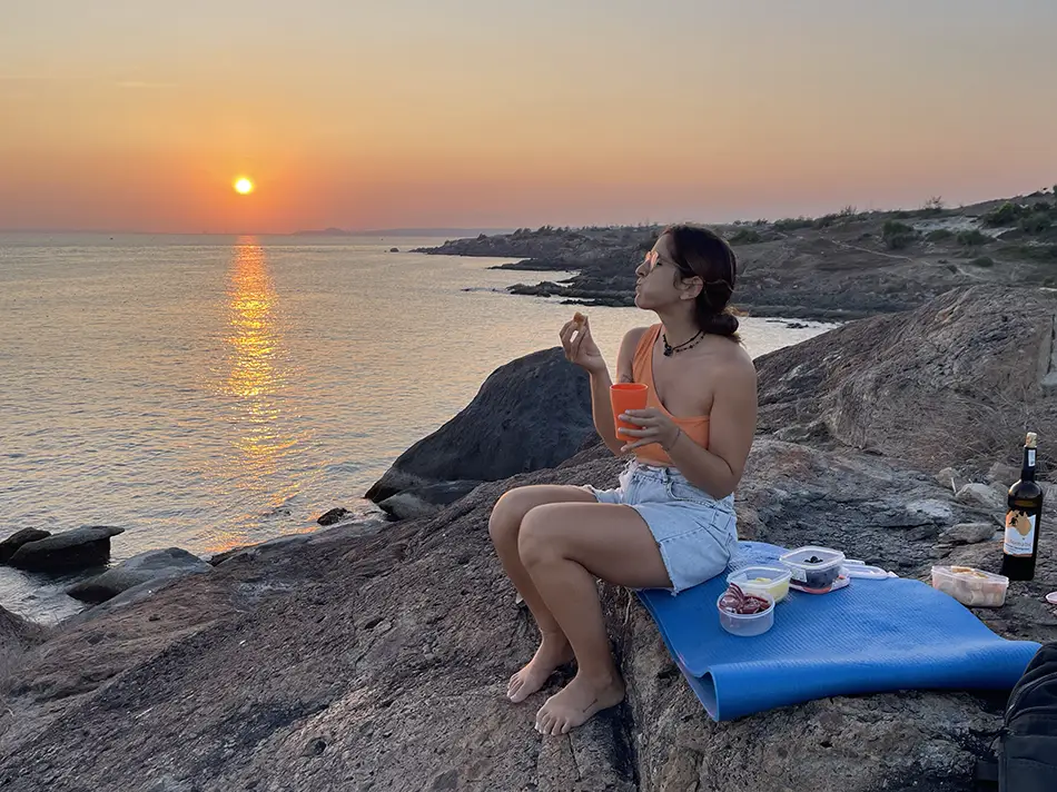 A traveler seated on stone slabs at the tip of the Mui Ne cape watching the sun set over the sea with a small picnic spread