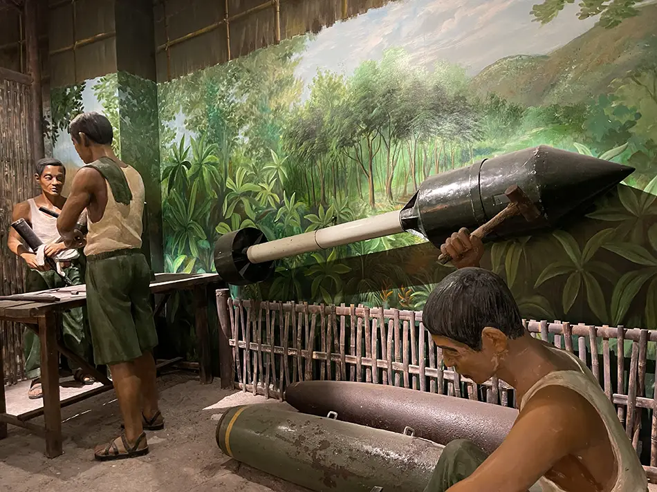 A visitor examining war-era weapons and military artifacts mounted against a painted jungle mural on the wall of a Vietnamese museum exhibition