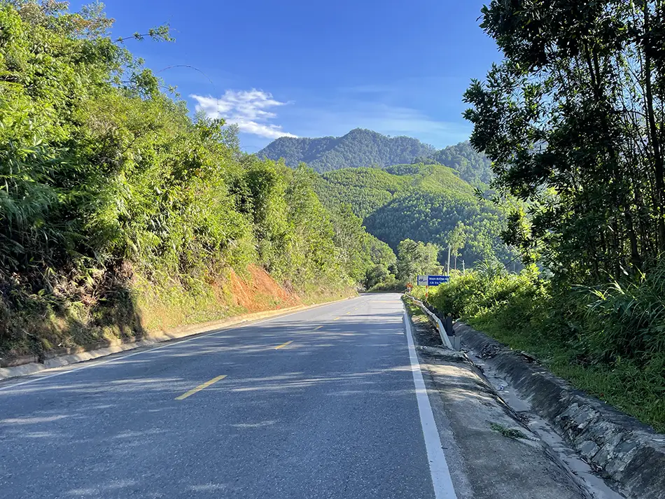 A road in Vietnam descending toward green hills surrounded by lush trees.
