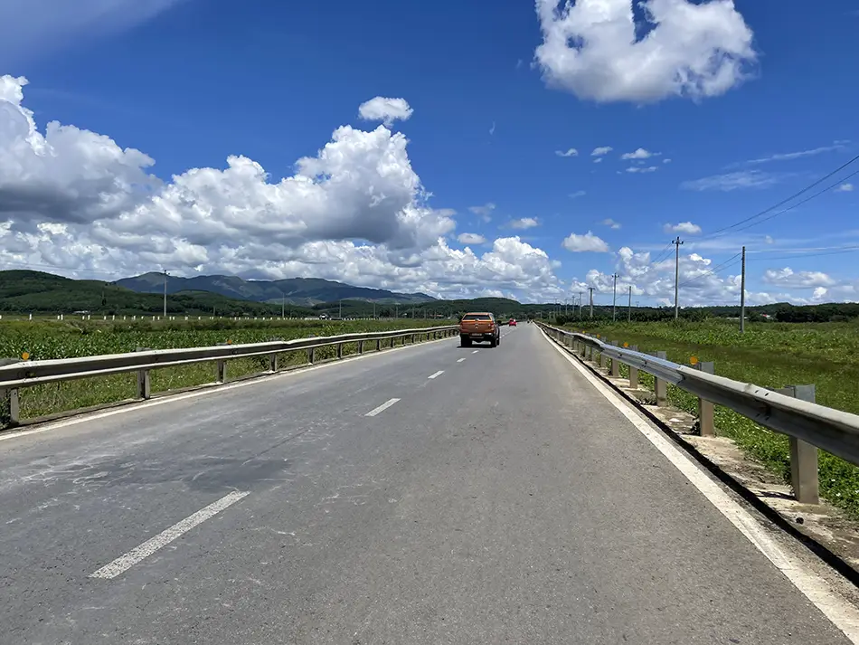 First-person view of a virtually empty road on a sunny day with green hills on the horizon.