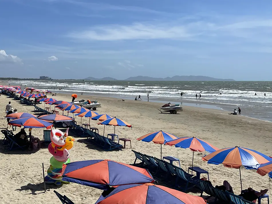 Rows of orange and red beach umbrellas with sunbeds lined up on a wide sandy beach, gentle waves and a clear blue sky stretching to the horizon