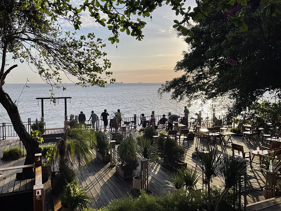 Wooden bench seating at a seaside café framed by tropical trees, with several visitors watching the sunset over the sea.