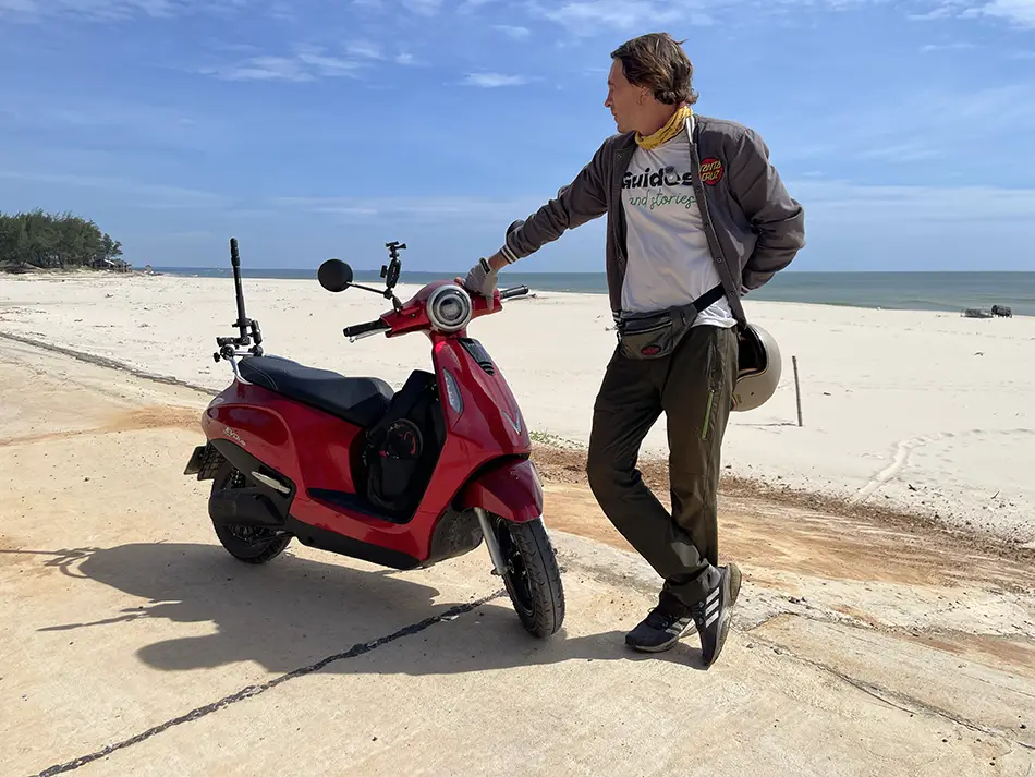 A traveler on a red electric scooter parked on a coastal sandy beach with the sea in the background.