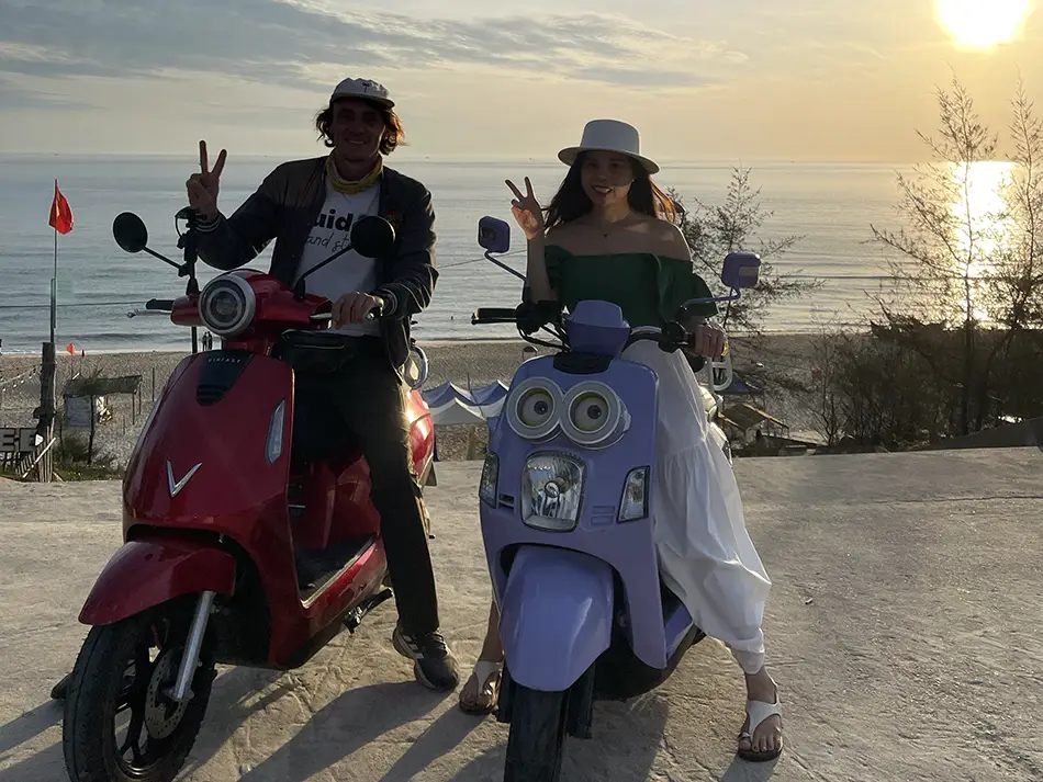 Two travelers on scooters at sunrise with the sea in the background.