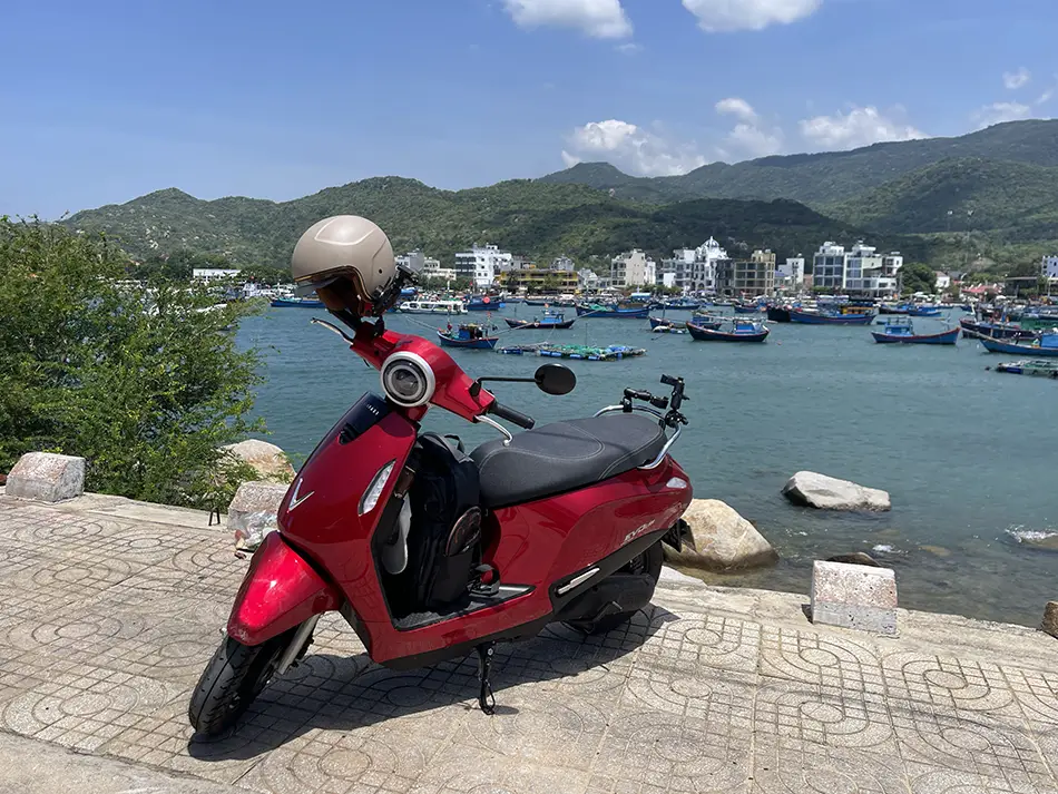 Red VinFast electric scooter parked above a harbor with boats and green hills in Vietnam.