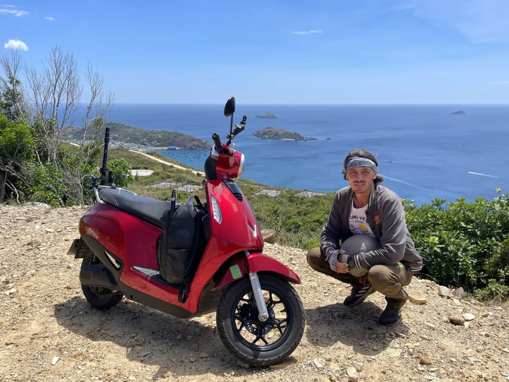 A traveler standing next to a red scooter at a lookout point with the sea stretching across the background.
