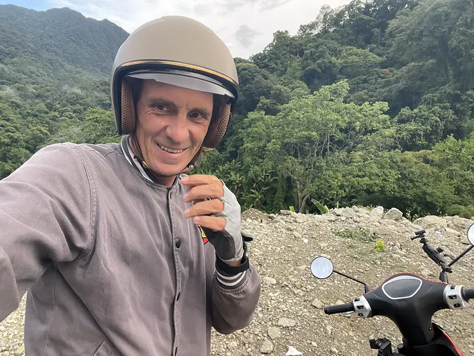 A traveler wearing a helmet next to a motorcycle on the roadside in Vietnam with jungle in the background.